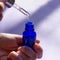 Square format of bottle with face skin care serum on purple background with long shadows. Female hands hold pipette with cosmetic