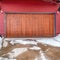 Square frame Brown garage door and red wall of home with snowy yard and driveway in winter