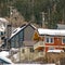 Square Mountain homes with snow covered roofs aginst coniferous trees and cloudy sky