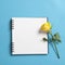 Square notepad on springs with white kraft paper with a yellow rose lay on a blue background.