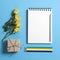 Square notepad on springs with white kraft paper with a yellow rose lay on a blue background. Copyspace