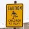 Square Yellow Caution Children At Play sign against blurry building and cloudy sky