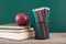 A stack of books with a red apple and a pen holder full of colored pencils in front of the blackboard