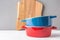 Stack of multicolored red and blue ceramic cooking pots wood cutting board on white table gray wall background. Food preparation