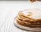 Stack Russian homemade thin pancakes on white plate and honey flows from wooden spoon. Traditional wheat pancakes for Shrovetide