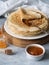 Stack Russian homemade yeast pancakes on white plate, wood honey spoon, honey in jars and apricot jam in bowl on grey table.