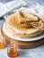 Stack Russian homemade yeast pancakes on white plate, wood honey spoon and honey in jars on grey table. Traditional wheat pancakes