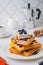 Stacked pumpkin waffles, sprinkled with powdered sugar, with fresh blueberries on a white plate on a light concrete background.