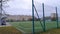 The stadium is located among trees, buildings and is surrounded by a mesh fence with metal posts. The soccer field is covered with