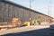 Staging area of road construction materials beside a concrete retaining wall and elevated highway