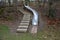 Stainless steel slide on the playground with concrete hills and rocks and wooden staircase. Large glossy tube for sliding children