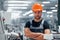 Stands with arms crossed. Portrait of male industrial worker indoors in factory. Young technician with orange hard hat