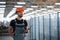 Stands in the corridor with documents in hands. Industrial worker indoors in factory. Young technician with orange hard hat