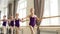 Starting ballet-dancers are doing exercises at ballet barre in spacious ballroom, their teacher in leotard is helping