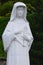 Statue of polish Saint Faustyna Kowalska holding cross and rosary, touching her heart, decorative shrubs in background.