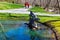 A statue of two boys sitting on a tree in a lake surrounded by silky green lake water, lush green grass, people walking