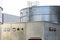 Steel industrial silos for liquids and solids standing in a factory next to a concrete building.