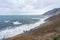 Steep cliffside with volcanic beach and waves crashing to shore. Mountains in the background.