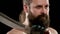 Stern man with beard, tattoo in T-shirt in black studio holding cleaver. Closeup