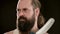 Stern man with beard, tattoo in T-shirt in black studio holding cleaver. Closeup