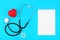 Stethoscope with blank notepad, red heart and white capsules on a blue background.