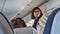 Stewardess in glasses looks toward back of plane viewed over heads of sitting passengers