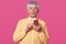 Stidio shot of mature white haired man dressed in yellow shirt and white bow tie, holding smartphone in hands and types message,