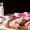 Still life with french sugared croissants, glass of milk and bowl of jam on old wooden board and black background