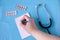 Still life : Notepad, tablets and stethoscope on a blue background, a hand writes a recipe in a notebook