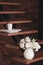 Still life of a vase with artificial white flowers and a stack of books with a cup stand on the steps of a wooden staircase