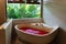 Stone bath tub with heart shaped flower petals near window with jungle view. Organic spa relaxation in luxury Bali