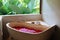 Stone bath tub with heart shaped flower petals near window with jungle view. Organic spa relaxation in luxury Bali bathroom
