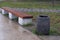 Stone black trash can with white splashes stands near brown bench on white blocks-legs near concrete walking area wet from rain