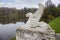 Stone concrete lion with wings statue, gargoyle on stone pedestal, shore of pond lake autumn landscape