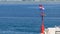 Stone pier with lighthouse and flying flag in daylight, people walking and sitting on stone wall, zoom out