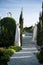 Stone walkway. Old statues of wife, women, Mary surrounds. Alley in beautiful garden with flowers and trees around. Summer in the