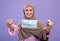 Stop covid concept. Excited muslim woman holding medical face mask in hands and smiling at camera, violet background