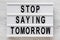 `Stop saying tomorrow` words on a lightbox on a white wooden background, top view. Overhead, from above, flat lay. Close-up