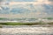 Stormy seascape with cloudy sky and waving sea surf on wet brown sand. Green sea water with white foam waves and dark