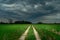 A straight dirt road through green farmland and dark storm clouds