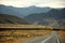 A straight two-lane asphalt road descends into a lowland surrounded by high, flat mountain peaks