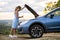 Stranded young woman driver standing near a broken car with popped up bonnet inspecting her vehicle motor