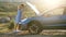 Stranded young woman driver standing near a broken car with popped up bonnet inspecting her vehicle motor.