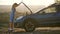 Stranded young woman driver standing near a broken car with popped up bonnet inspecting her vehicle motor.