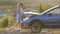 Stranded young woman driver standing near a broken car with popped up bonnet inspecting her vehicle motor.