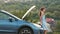 Stranded young woman driver standing near a broken car with popped up bonnet inspecting her vehicle motor