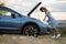 Stranded young woman driver standing near a broken car with popped up bonnet inspecting her vehicle motor