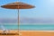 A straw umbrella and a white chair stand on a sandy beach near the water. Rest on the Dead Sea in Israel overlooking the mountains