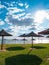 Straw umbrellas on green grass harsh shadow, sand beach vivid blue sea water sky background sunbeam. Scenic landscape