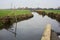 Stream of water in the countryside and a mooring pole by the shore with its reflection casted in the water on a cloudy day